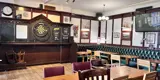 A room in a pub. There is a dart board at the centre of the image, with chairs and tables in the foreground. There is dark green cushioned seating on the right 