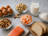 An image of a variety of food allergens. in a clockwise direction starting from the top left there are 6 eggs in an egg box, a glass bowl of peanuts in shells, a glass of milk, a glass bowl of flour, 3 slices of bread on a wooden chopping board, a piece of raw salmon on a paper towel on a wooden block, a bowl of mixed tree nuts and then in the centre is a glass bowl of cooked prawns. They are all placed on a pale grey surface.