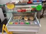 a deli fridge containing a variety of sandwich fillings in sealed tubs at the front of the lower shelf. To the rear of the lower shelf on the left are some small pies and to the right are some larger sized pies. On the upper shelf there is a variety of chocolate bars. On the top left of the fridge there is a yellow sign and on the right is a bright green sign with promotion details and product details