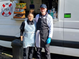 The two owners of Canal side Cafe stood in front of their food truck. The person on the left is female and the person on the right is male. 