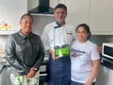 Three people smiling towards the camera. They are stood in a home kitchen. From left to right there is a female, a male and a female. The male has his arm around the shoulder of the female to the right. The man is wearing a chefs outfit. The female on the left is wearing a black leather jacket and a flower print knee length dress and the female to the rights is wearing a white reebok branded tshirt. 