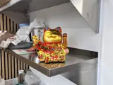 An image of a gold coloured Chinese style lucky waving cat on a metal shelf in a takeaway kitchen