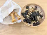A small white paper bag on the left with two bone shaped dog treats. Porridge in a cardboard pot on the right, topped with seeds and dried blueberries. 