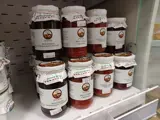 A variety of chutneys stacked on a white shop shelf. There is a white paper frill covering each jar. There is a small logo on a white label of each jar. 