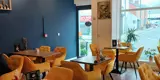 A coffee shop dining room with wooden tables, yellow padded chairs and dark blue walls. There are three lights hanging from the ceiling and a wooden floor
