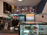 A man with brown hair stood behind a coffee shop counter. To the right of the counter is a glass containing cakes. At the top of the image is a menu board