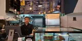 A man with brown hair stood behind a coffee shop counter. To the right of the counter is a glass containing cakes. At the top of the image is a menu board