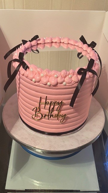 A pale pink birthday cake with a mirrored top. There are also small white pearl decorations and four thin black ribbon bows on the piped border on the top edge of the cake. There is a golden "happy birthday" decoration on the front, middle section of the cake.