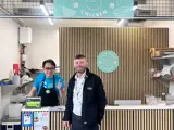 Two men stood smiling. One of the men is in front of a till and one is behind the till. The man behind the till has his thumb up