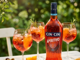 A bottle of premium alcohol-free spirit style drink on a table in a garden. The drink is deep orange in colour. There are also three wine glasses filled with the beverage on the table and some pieces of orange for decoration, along with a shallow dish of olives. 