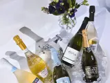A variety of sparkling alcohol free bottles of wine artistically places in a white sink of ice. There is a large wine glass containing some dark purple flowers with dark green leaves above the sink. 