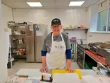 The owner of Penny's Pantry - a lady smiling towards the camera while stood in a professional kitchen. The lady is wearing a white apron and a black peak cap. 