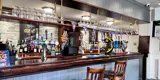 The bar in the Morris Dancers pub in Bolton. The bartop is dark wood and there are two stools at the bar in the middle of the image. Behind the bar there are beer pumps and there is a shelf full of drinking glasses above the bar